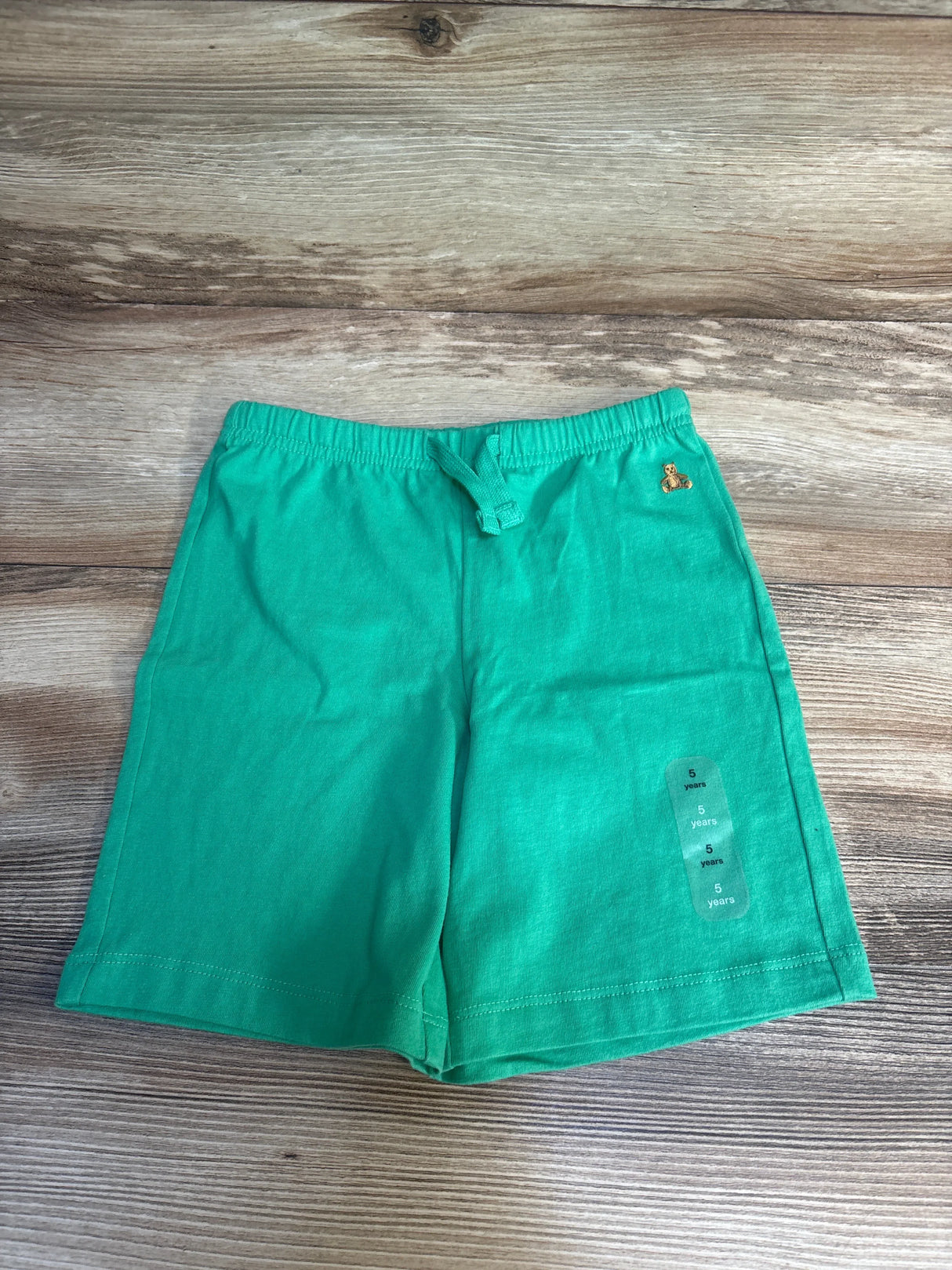 Green kids shorts with drawstring waist, teddy bear logo, size 5 years, laid on wood floor