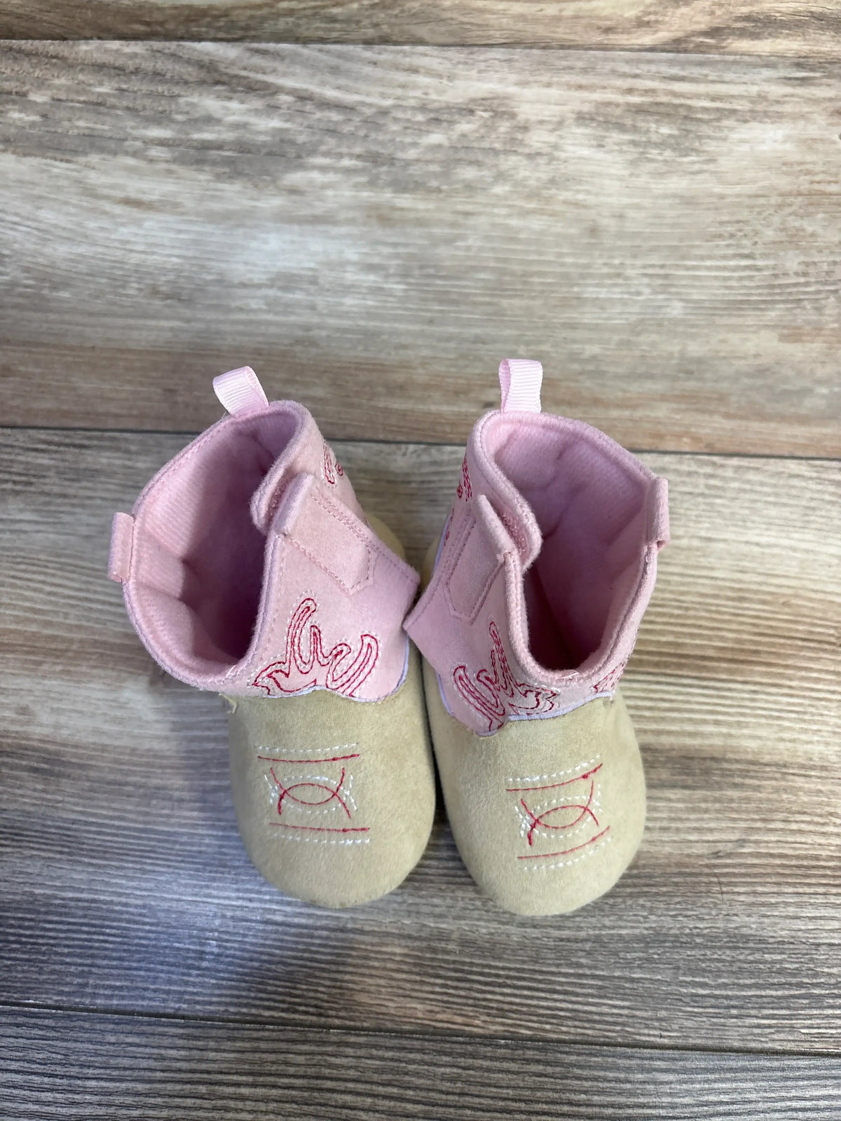 Top view of Stepping Stones cowgirl crib boots in pink and tan, showcasing embroidered Western designs on a wood background
