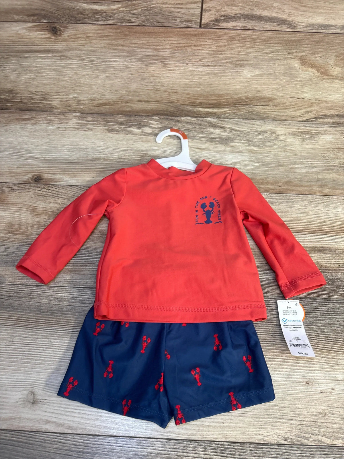 Red long sleeve baby rash guard and navy swim shorts with lobster print on wood floor.
