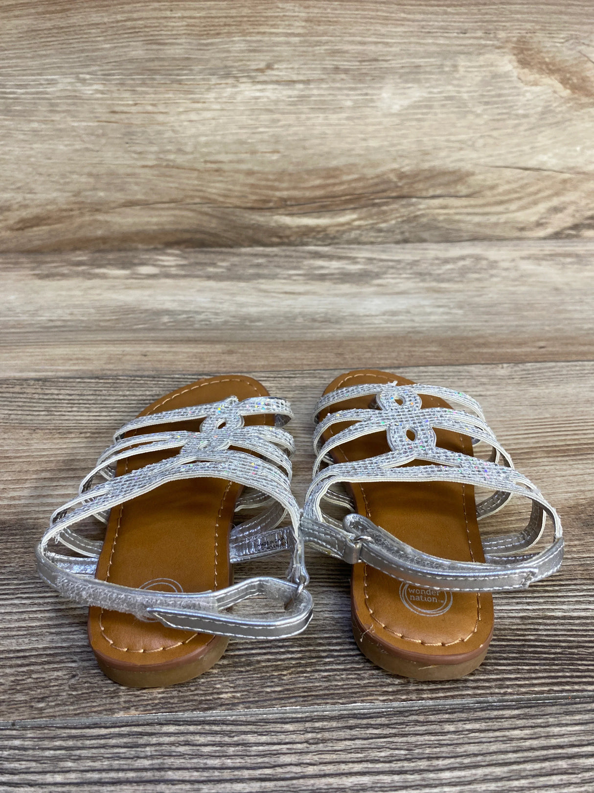 Girls' silver strappy sandals with flat brown soles on wooden background