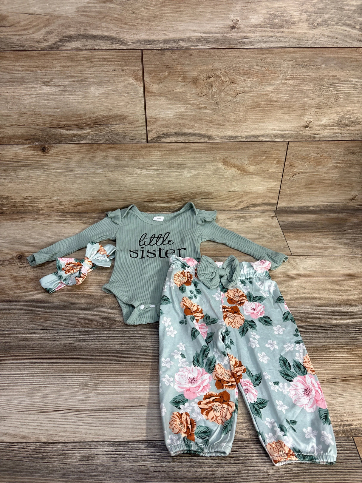Flat lay of green 4-piece baby outfit: ribbed onesie reading little sister, floral elastic-waist pants, matching headband
