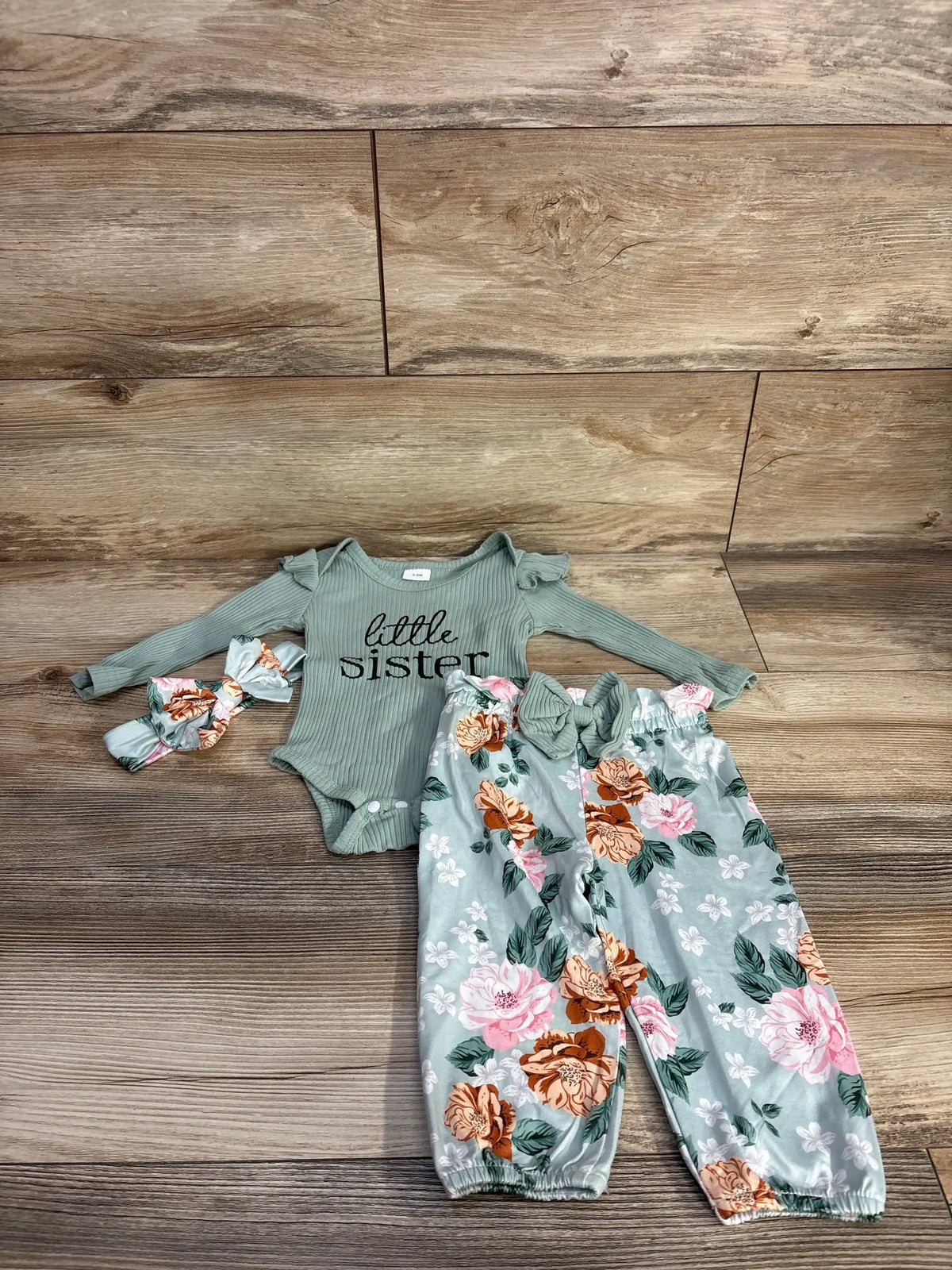 Flat lay of green 4-piece baby outfit: ribbed onesie reading little sister, floral elastic-waist pants, matching headband