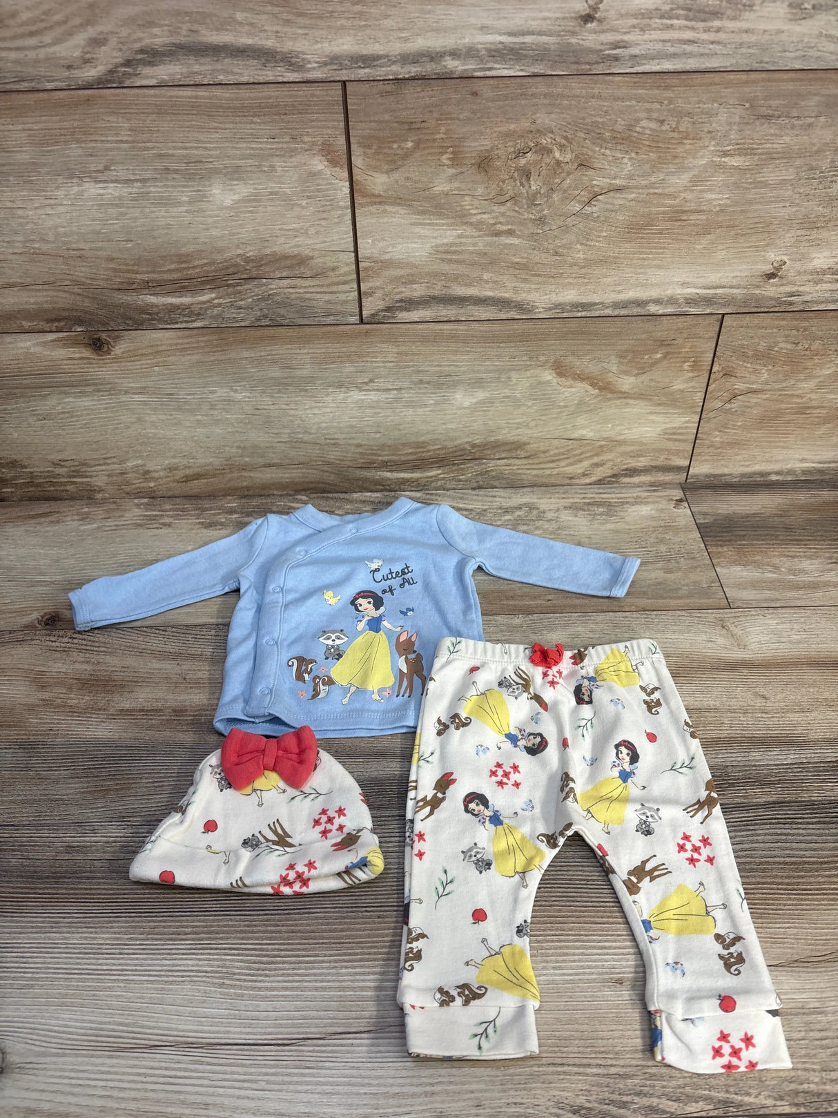 Flat lay of 3-piece Disney set: blue Snow White top 'Cutest of All', printed pants and hat with red bow on wood