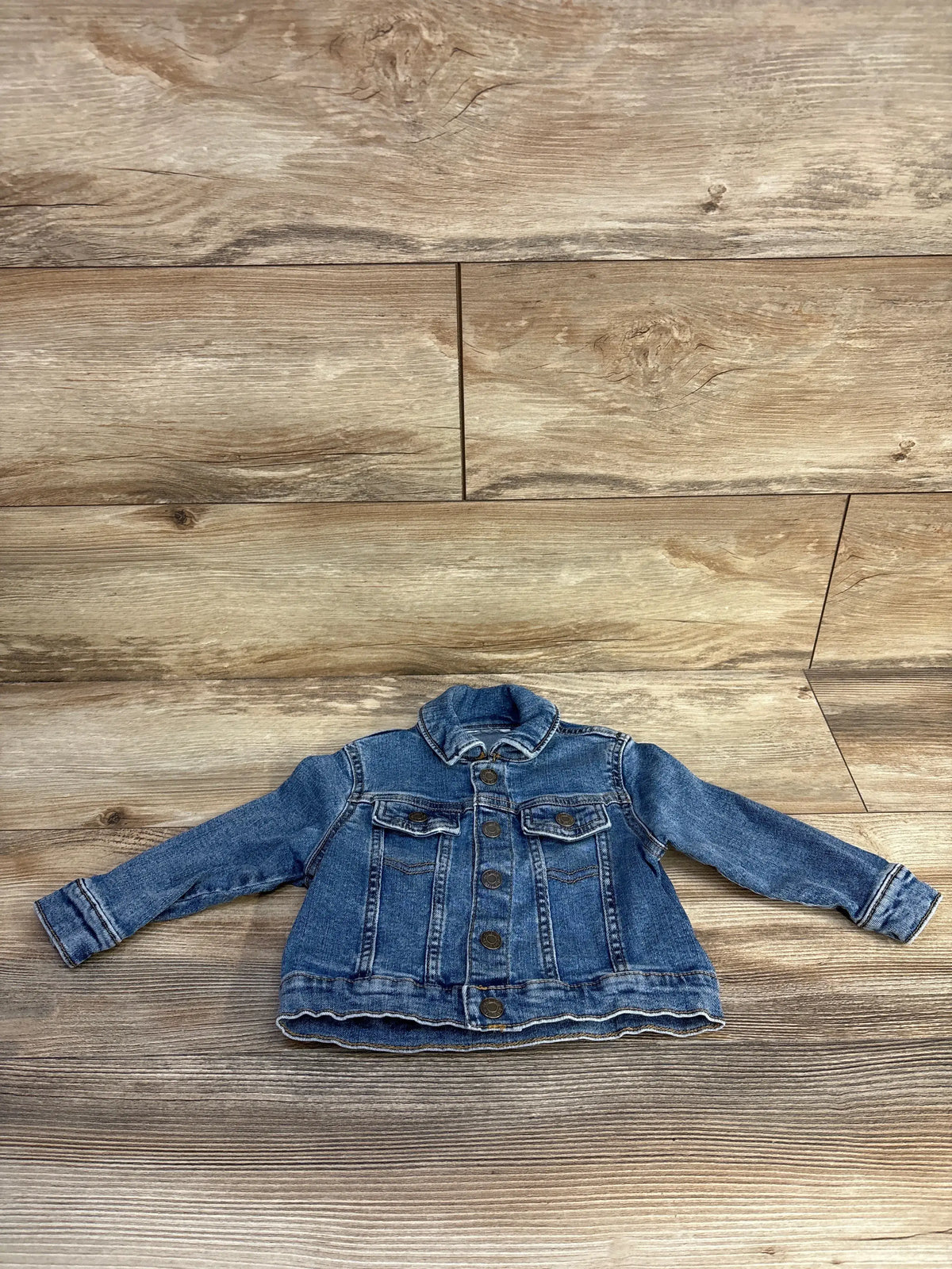 Front view of blue Okie Dokie toddler denim jacket showcasing button front long sleeves and chest pockets on a wood floor