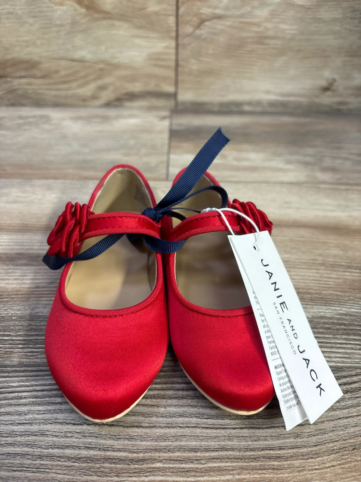 Front view of Janie and Jack red satin ballet flats showcasing rosette ankle straps with navy ribbon tie on wood floor
