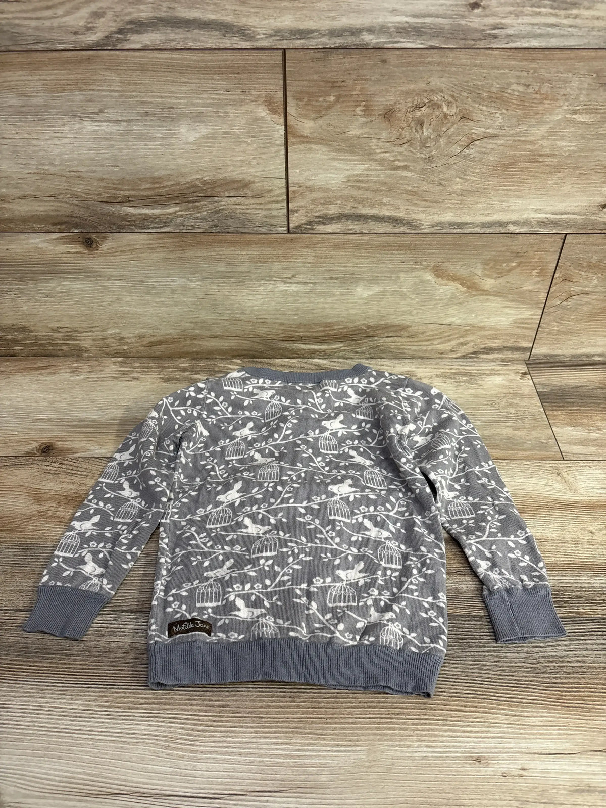 Gray long sleeve kids sweater with white bird and cage print, laid flat on wood floor