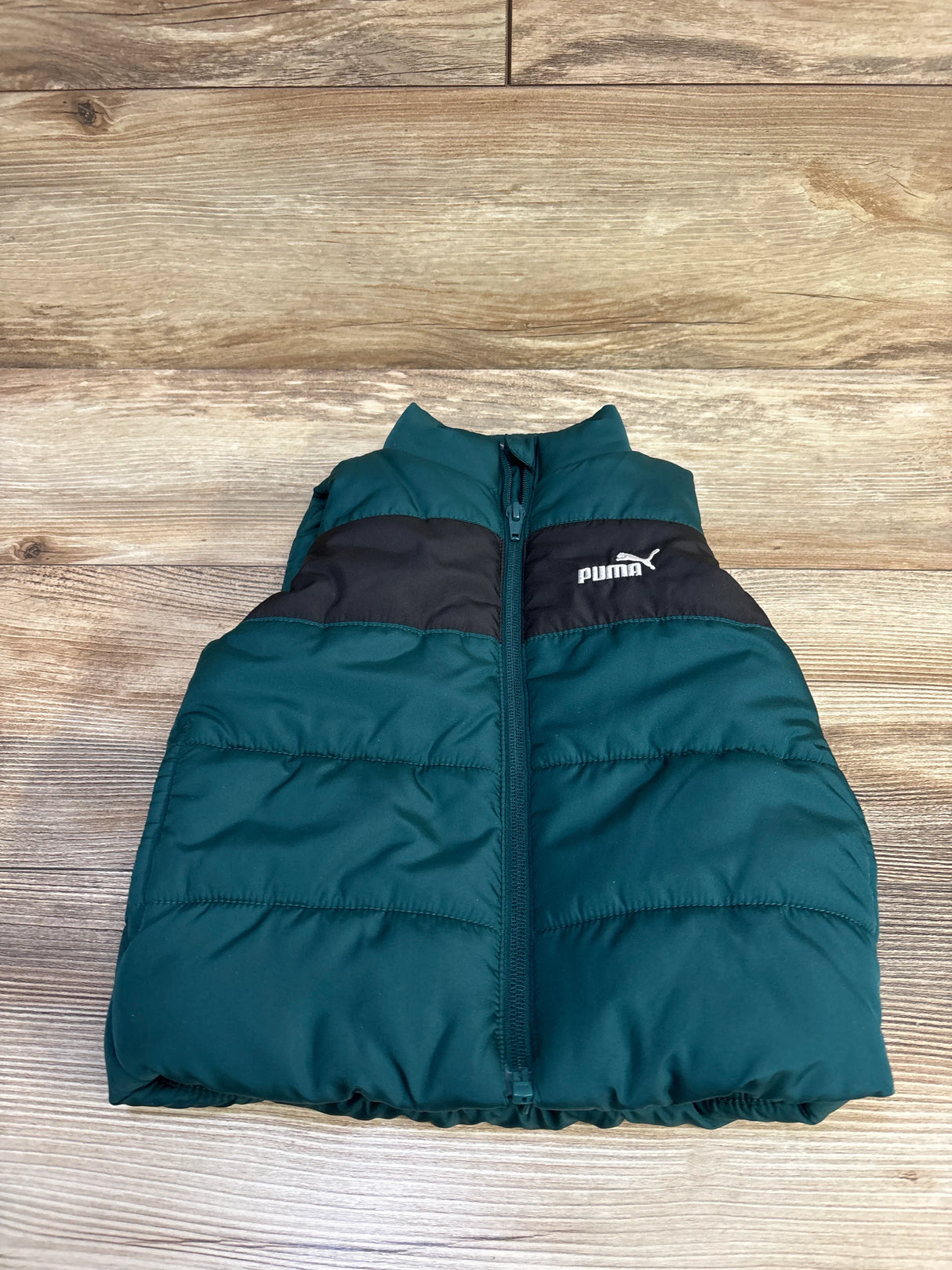 Front view of toddler Puma puffer vest in dark green with black chest panel and embroidered Puma logo, zipper closed on wood floor