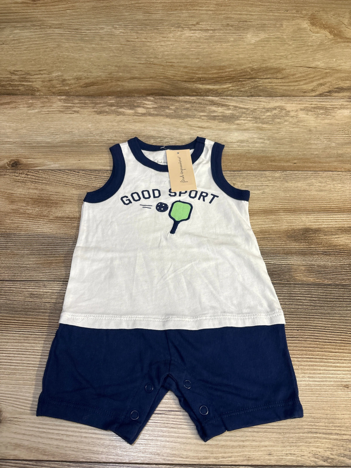 Flat lay of First Impressions romper, sleeveless white top with navy trim and Good Sport paddle graphic, navy shorts on wood background