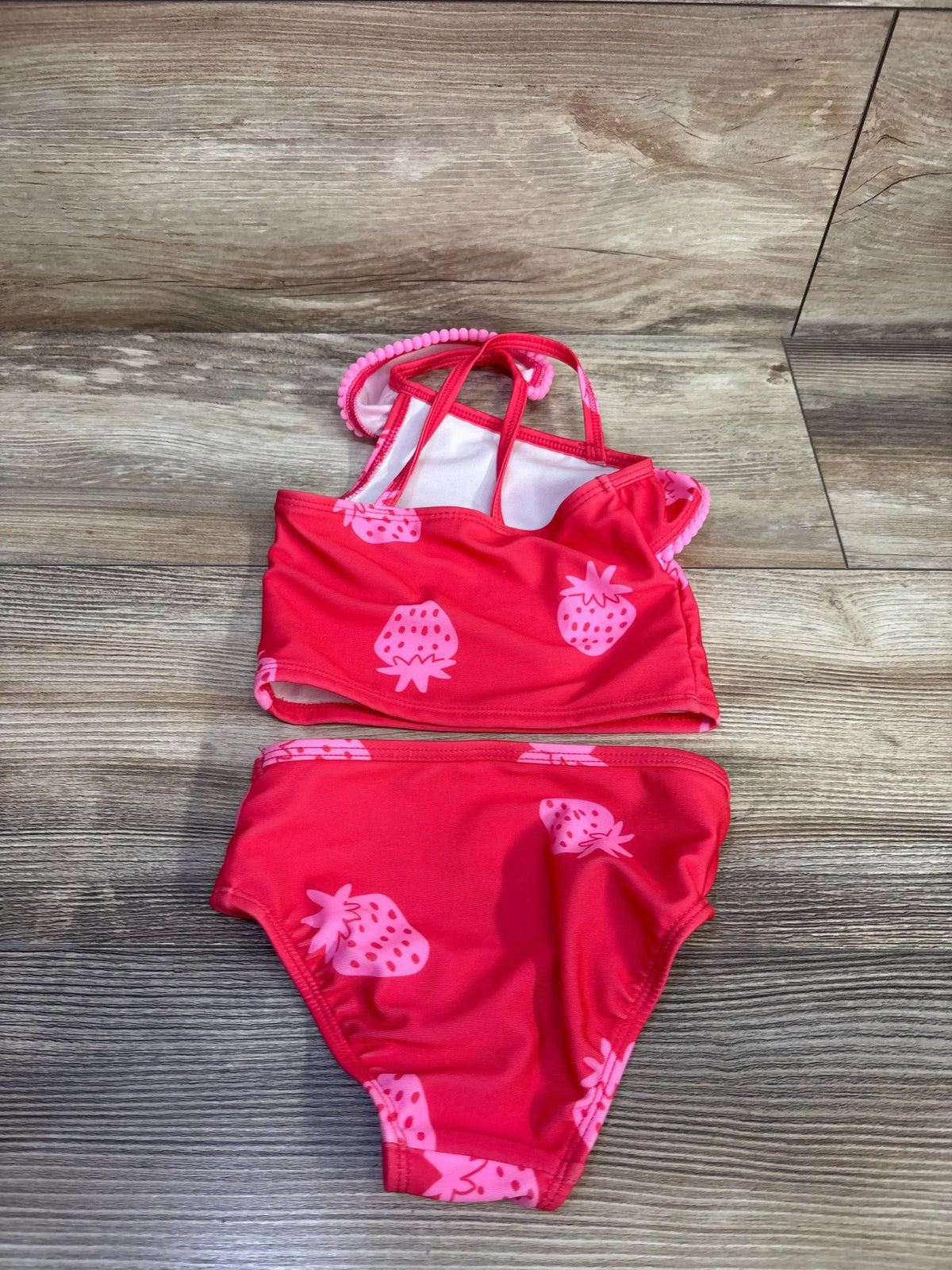 Flat lay of toddler two-piece red strawberry swimsuit size 12 months showing strawberry print and crisscross straps on wood