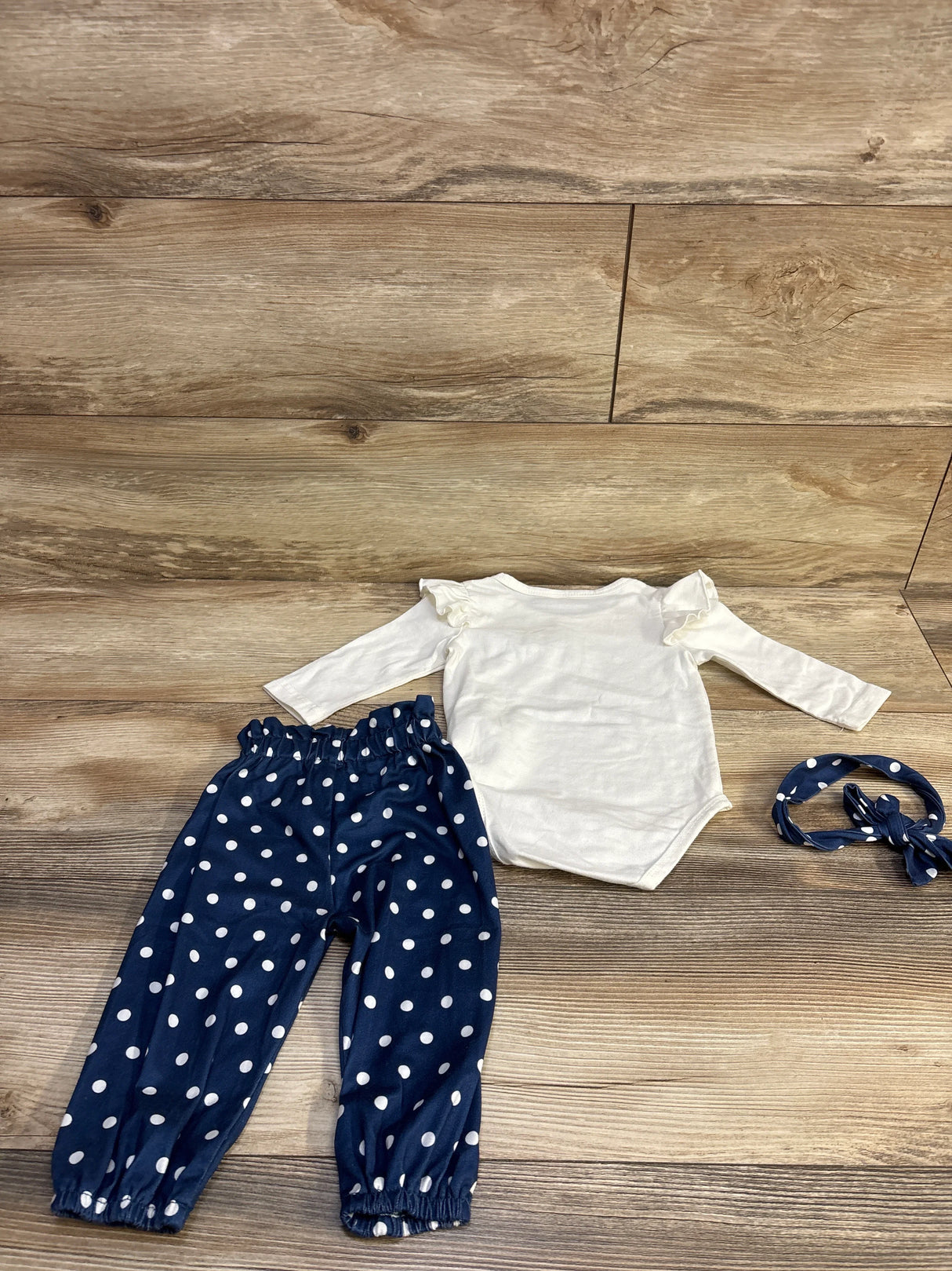 Flat lay of 3-piece baby set: white long-sleeve ruffle bodysuit, navy polka-dot pants, and matching headband on wood background