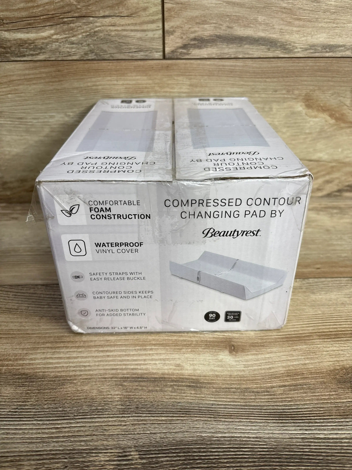 Front view of Beautyrest compressed contoured changing pad box showcasing waterproof vinyl cover, safety straps and product callouts on wooden surface