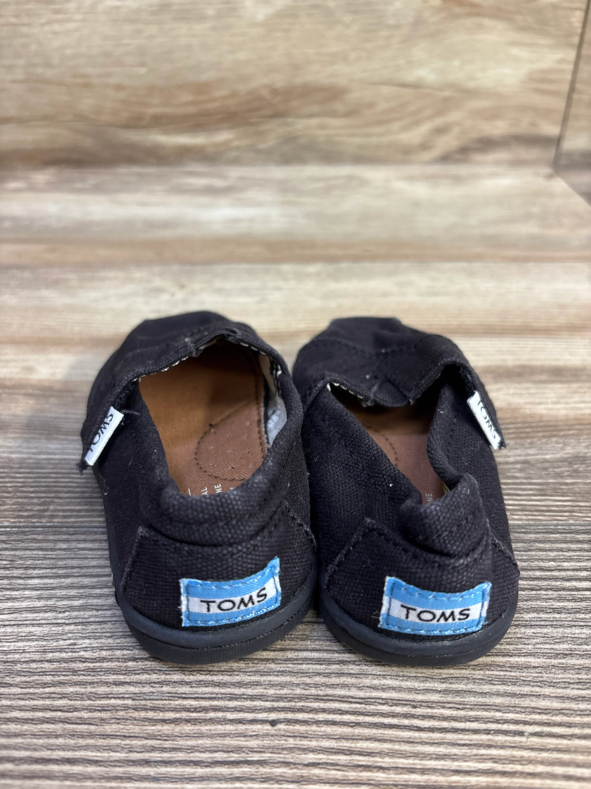 Rear view of toddler TOMS Tiny Alpargata shoes in black showing blue TOMS heel labels on a wood-grain surface
