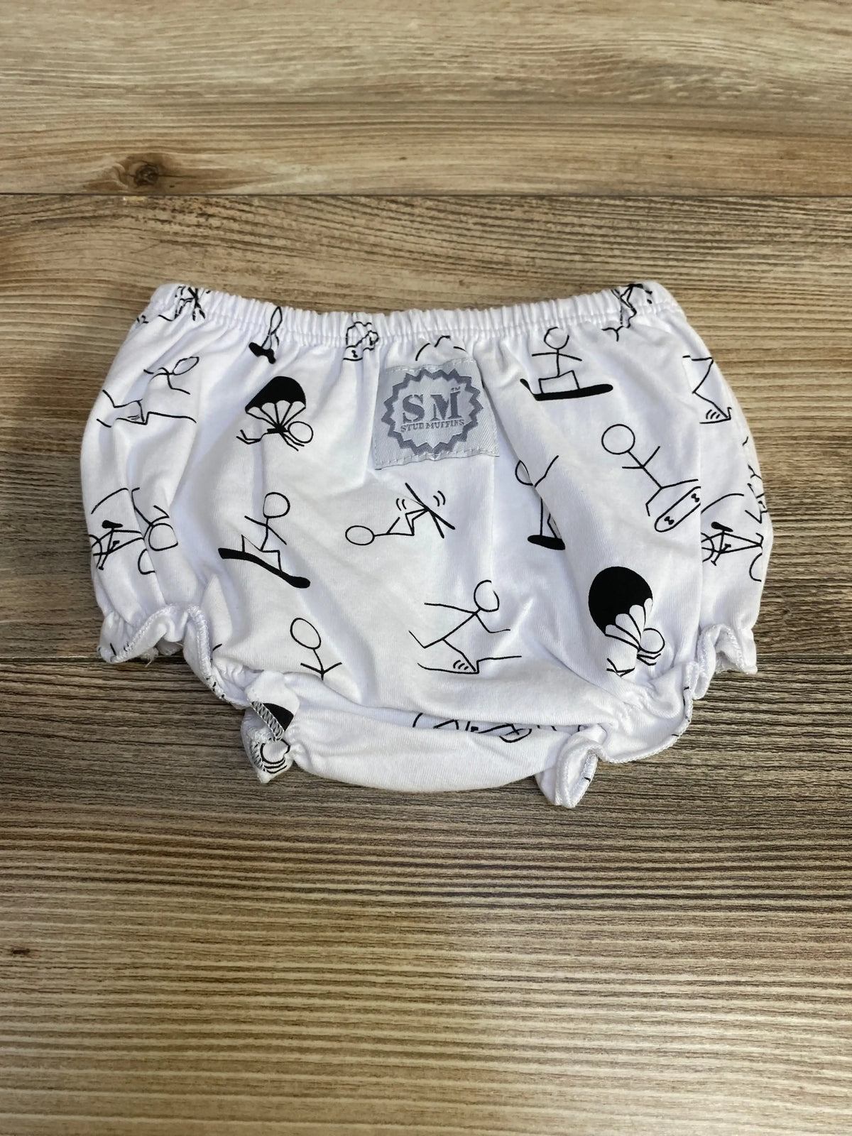 Top-down view of white Stud Muffins baby bloomers with black stick-figure print and elasticized waist on wood background