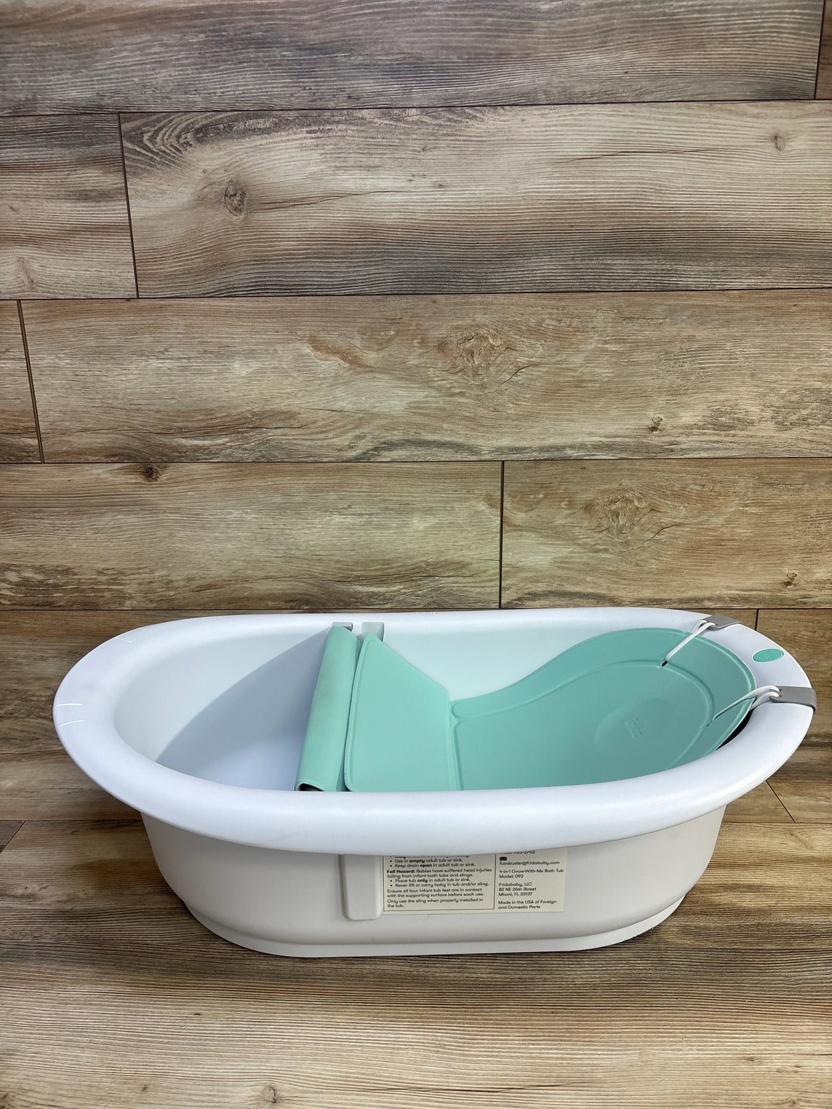 Angled view of Frida Baby 4-in-1 Grow-With-Me Bath Tub showing mint infant insert and white tub on wood background