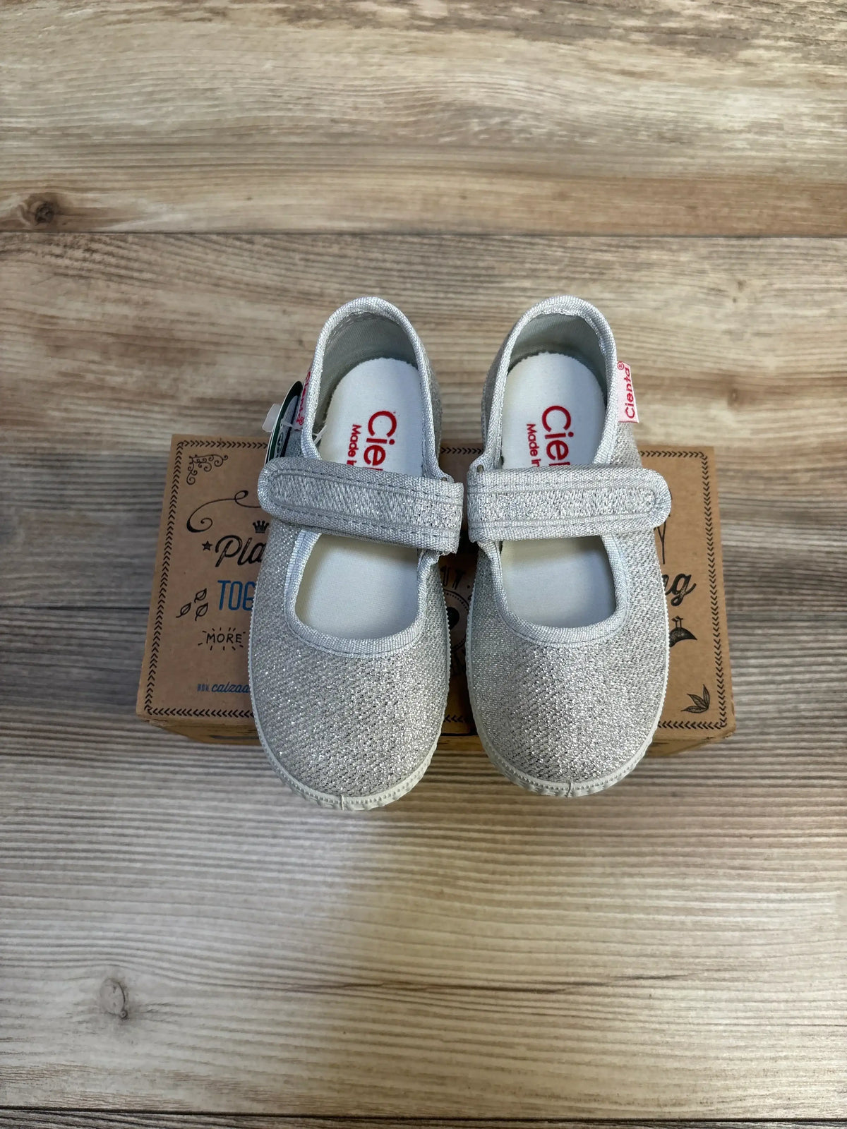 Top-down of Cienta Mary Jane toddler flats size 7C with silver glitter and Velcro strap on shoebox on wood background