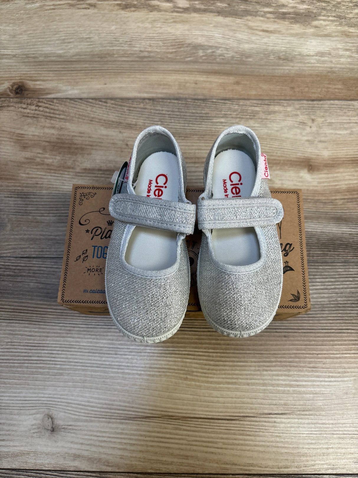 Top-down of Cienta Mary Jane toddler flats size 7C with silver glitter and Velcro strap on shoebox on wood background