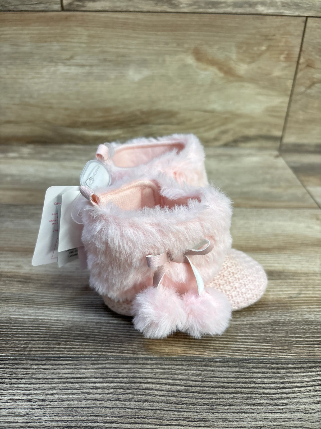 Side view of pink knit baby booties with faux-fur cuff and pom-pom bows on wooden plank background