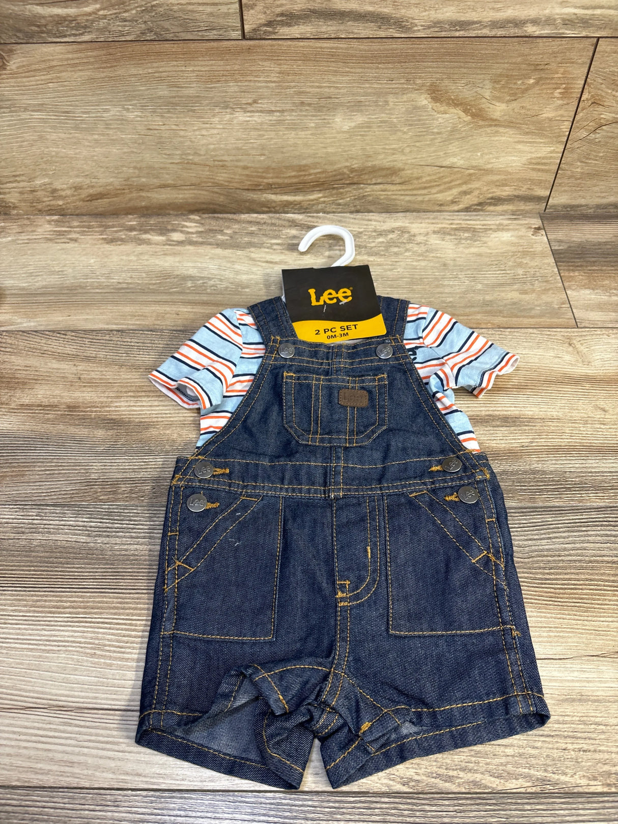Front flat lay of Lee newborn denim shortall with striped light blue tee showing front chest pocket and contrast stitching on wood background