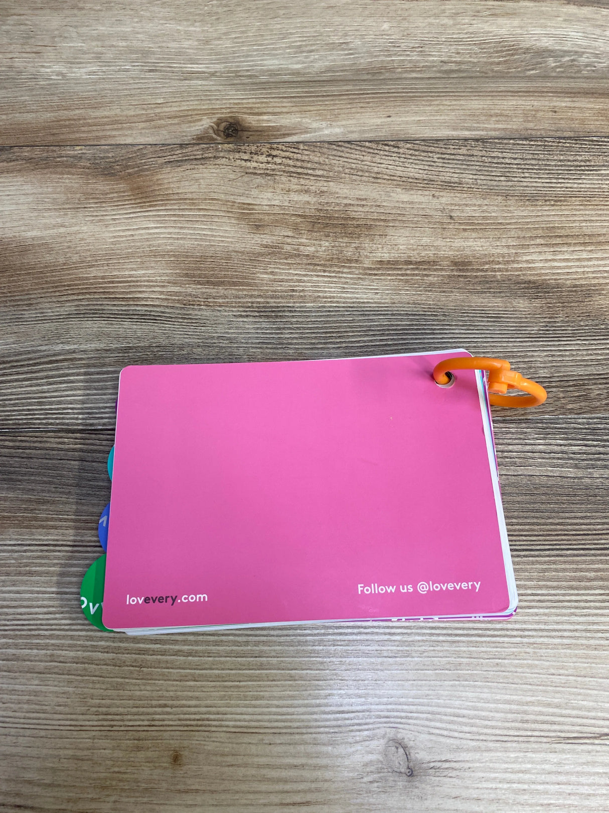 Pink card booklet with orange ring on wooden table, showing lovevery.com and social media handle