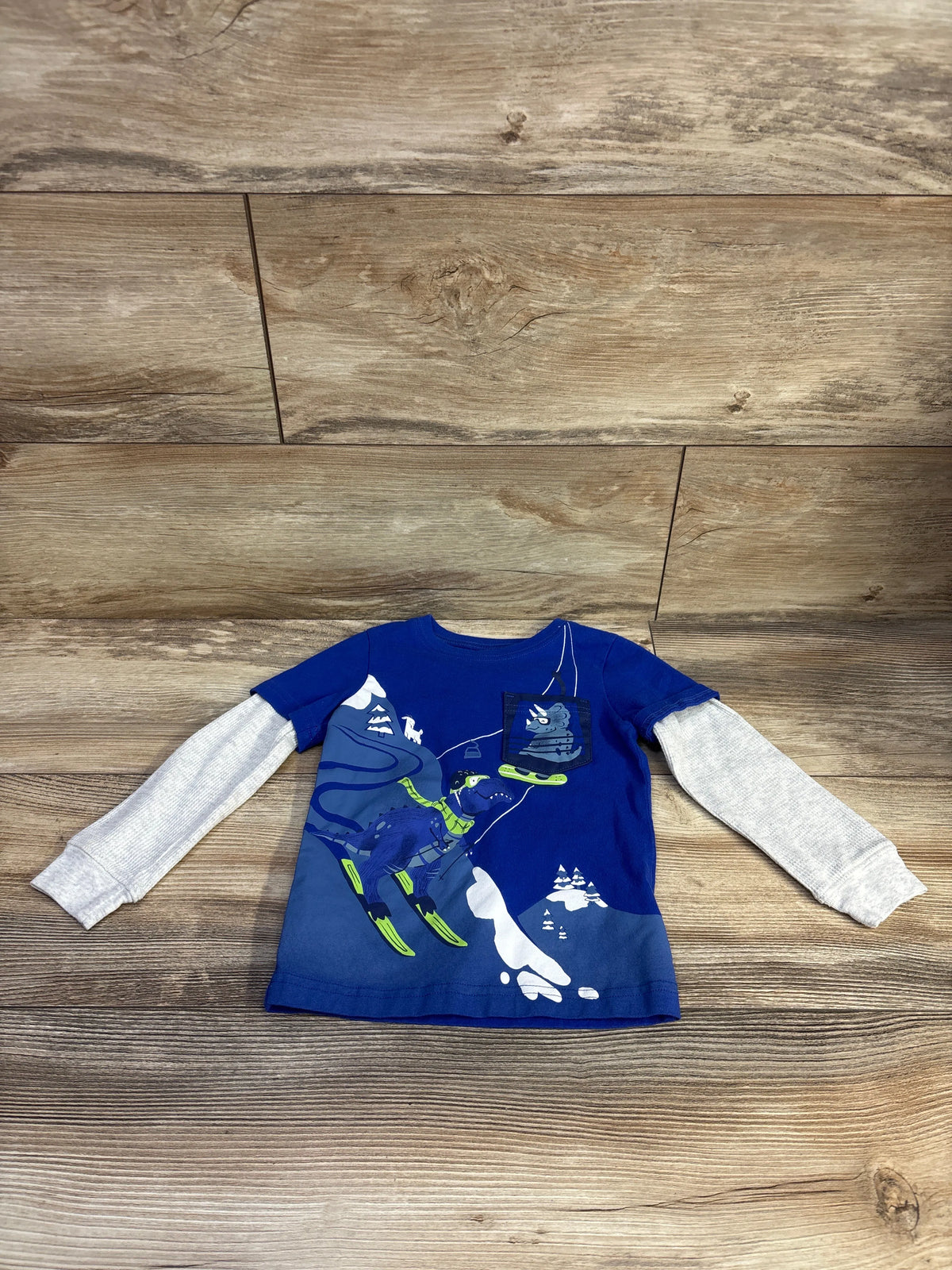 Flat lay of toddler blue layered-sleeve shirt showing gray long sleeves and neon-accent dinosaur skiing graphic on wood background