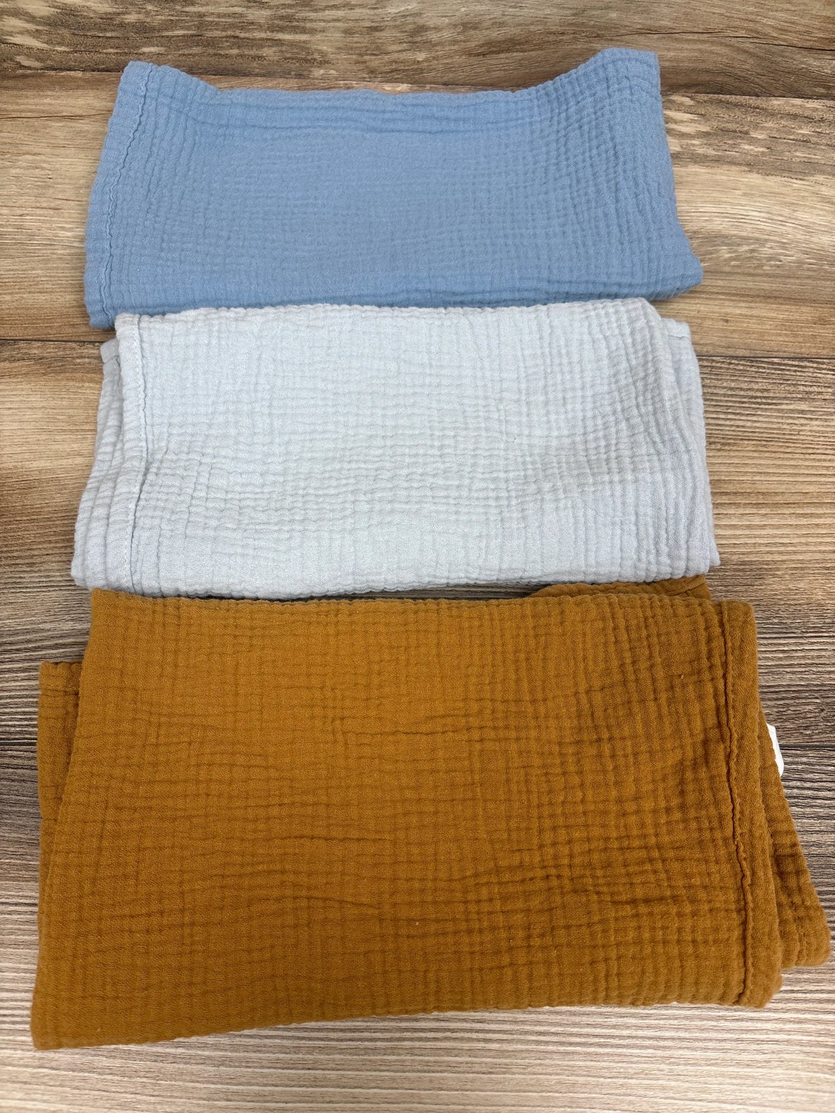 Top view of three folded 23 by 23 inch muslin security blankets and burp cloths in blue, white, and mustard on wood