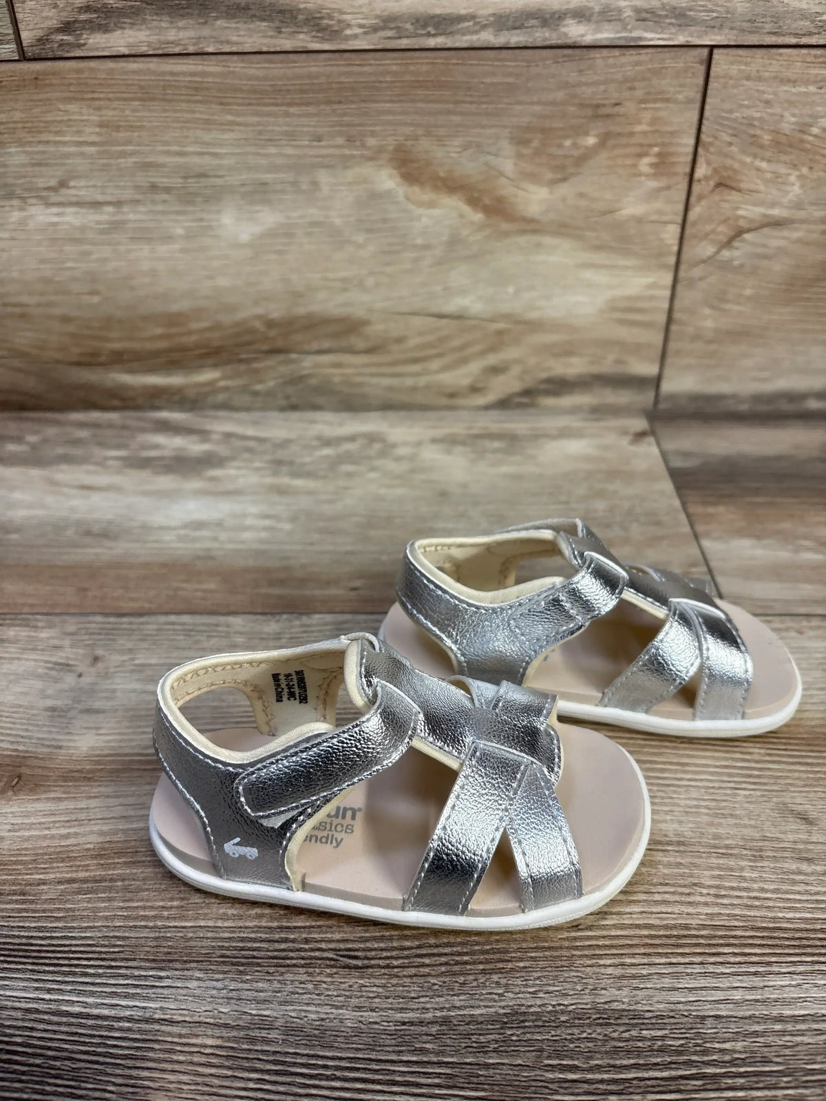 Front three-quarter view of See Kai Run Shayna sandals showcasing silver metallic strappy uppers and adjustable straps on a wood floor