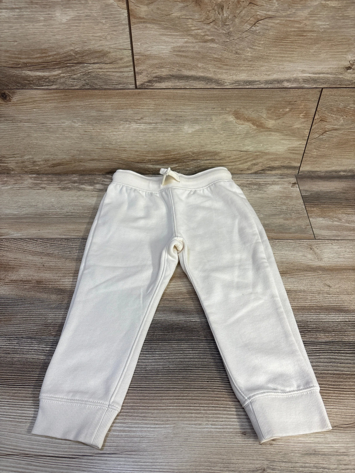 Front flat lay of cream Oshkosh fleece joggers size 3T with ribbed cuffs and drawstring waist on light wood background