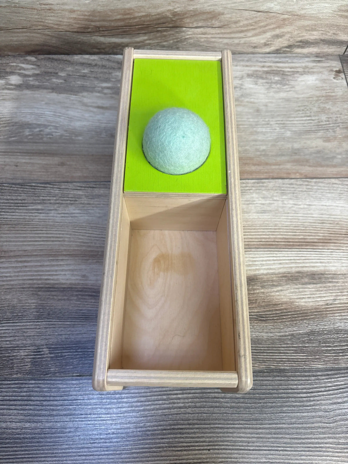 Top down view of Lovevery Sliding Top Box showcasing green sliding top and felt ball in compartment on wood grain surface