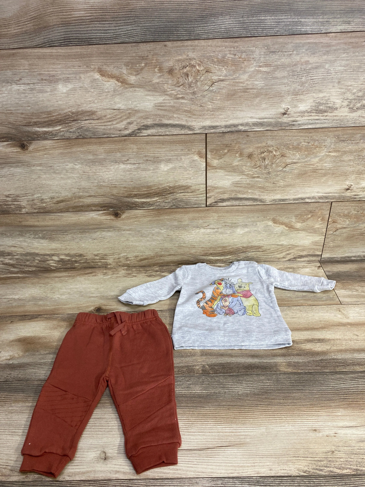 Toddler outfit with rust-colored pants and a gray long-sleeve shirt featuring Winnie the Pooh characters on wooden floor