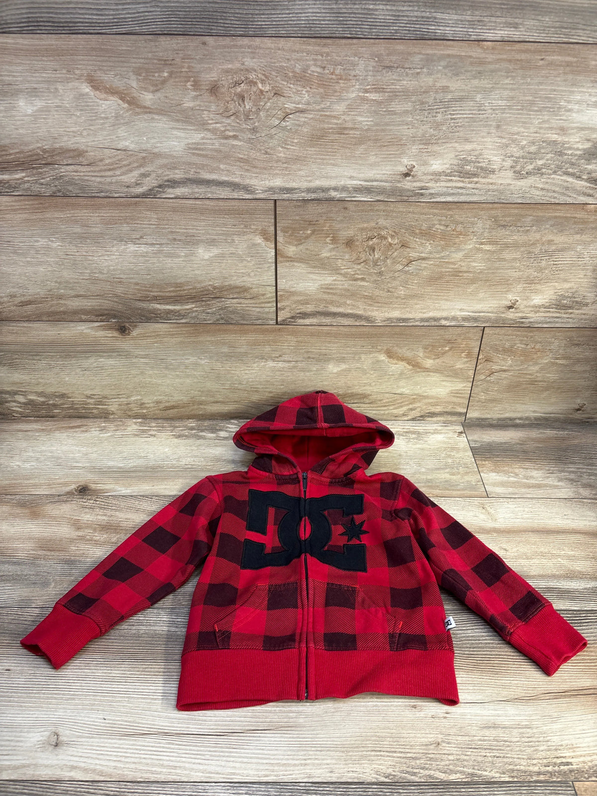 Front view of toddler red and black plaid DC Shoes zip-up hooded sweatshirt size 2T showcasing large black DC logo, front pockets, and ribbed cuffs on light wood background