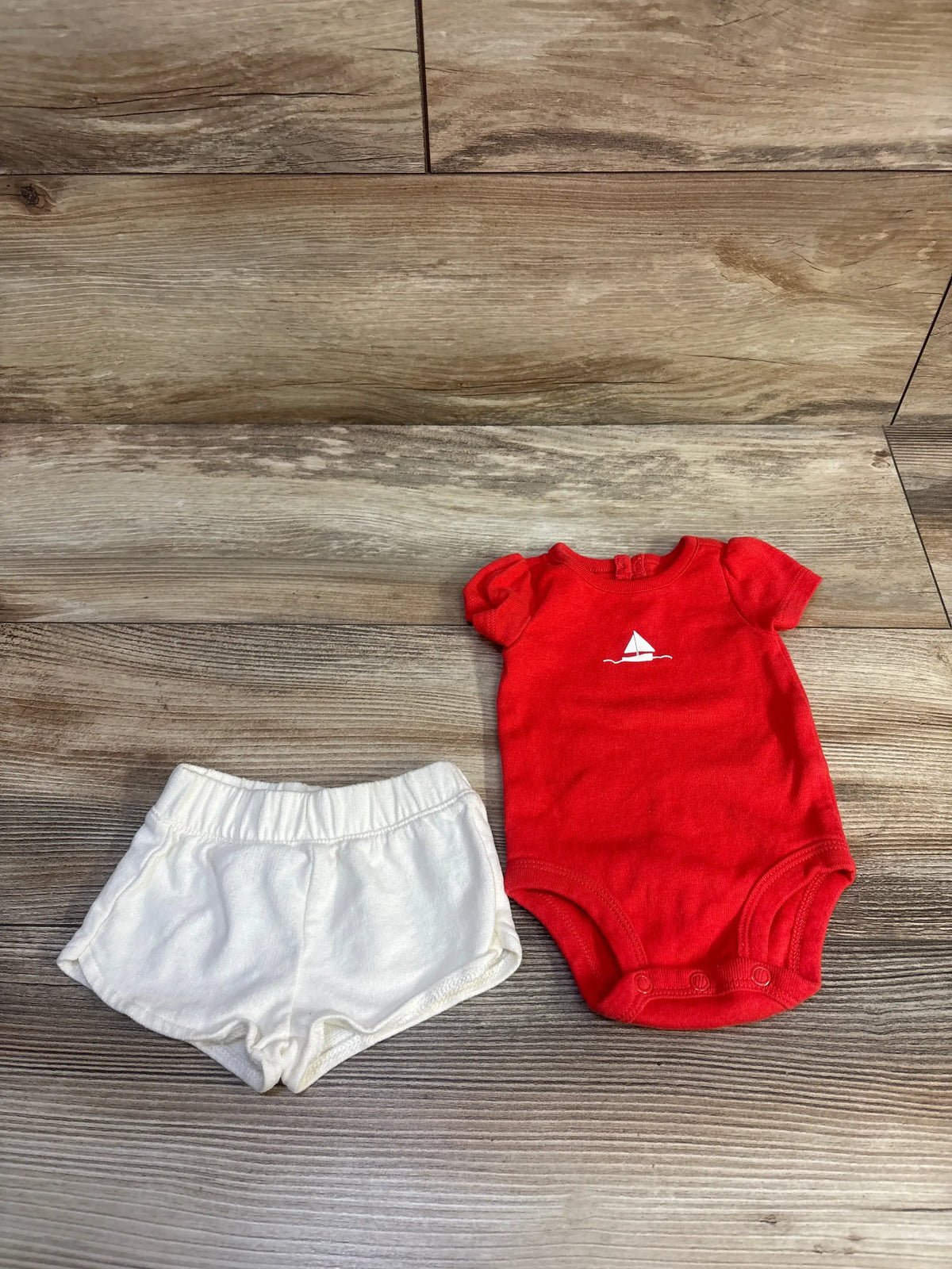 Top view of Carter’s 2pc Shirt and Shorts Set with red bodysuit, white sailboat graphic, and white shorts on wood floor