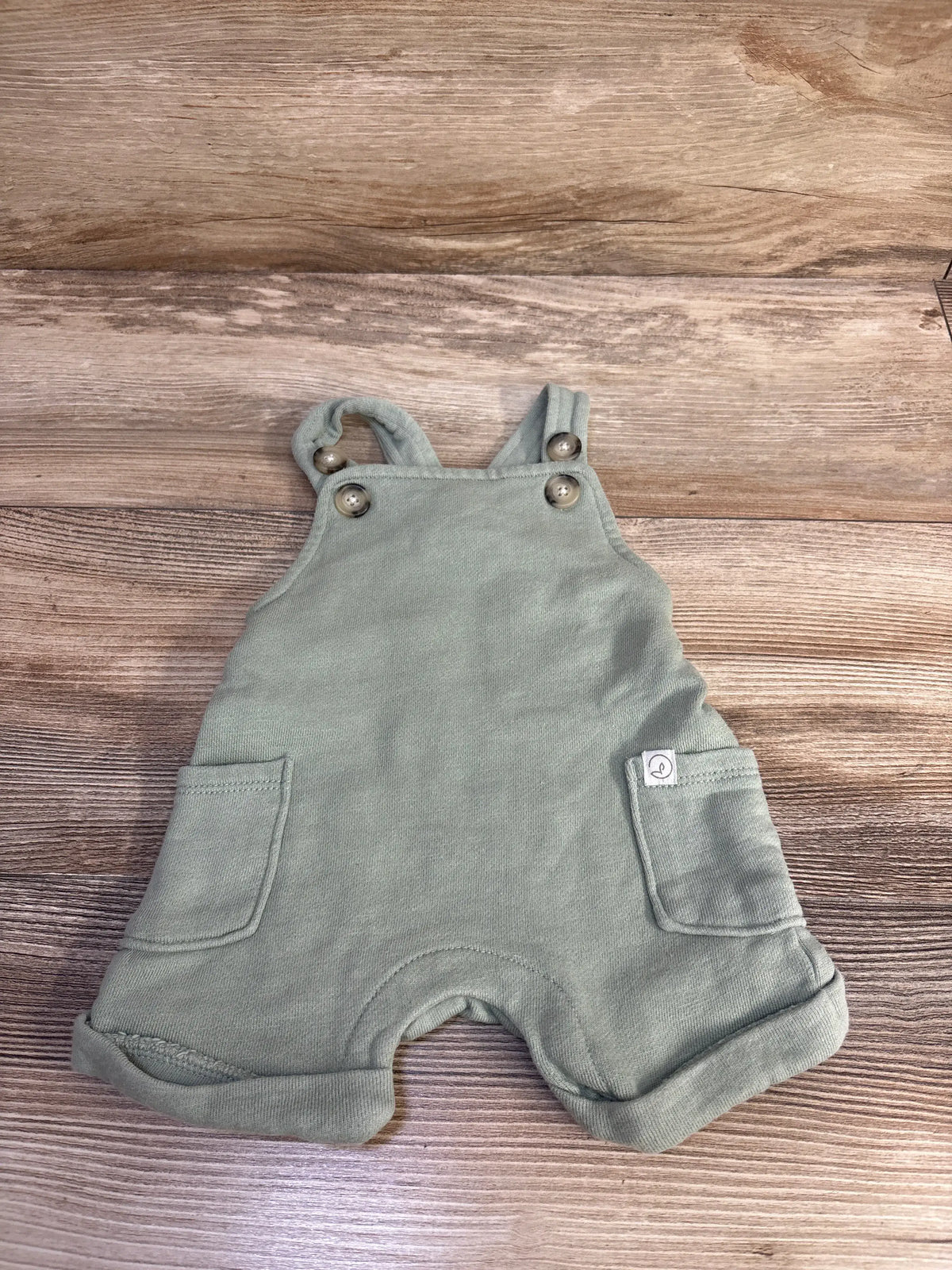 Front view of green organic shortalls size 3M with button straps, front pockets and rolled cuffs on wood background
