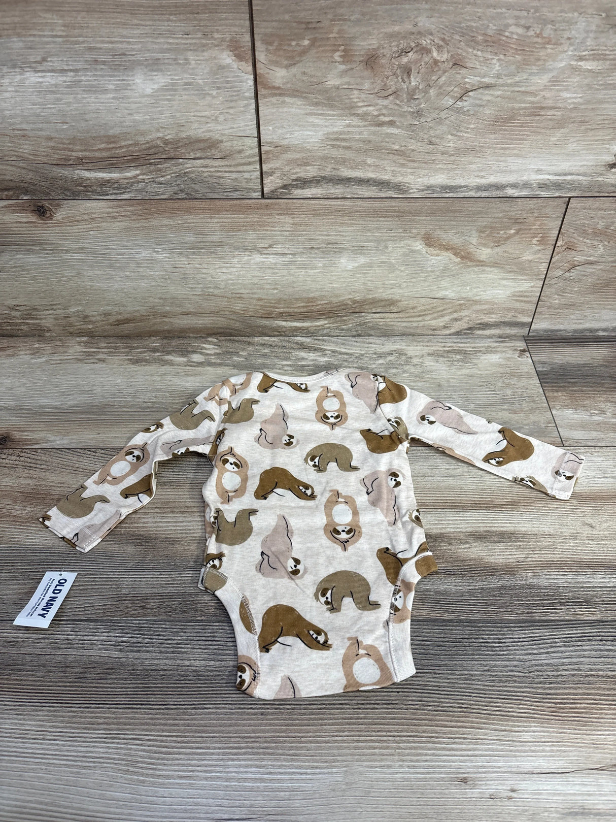 Beige long sleeve baby bodysuit with cute sloth print, laid flat on wood floor
