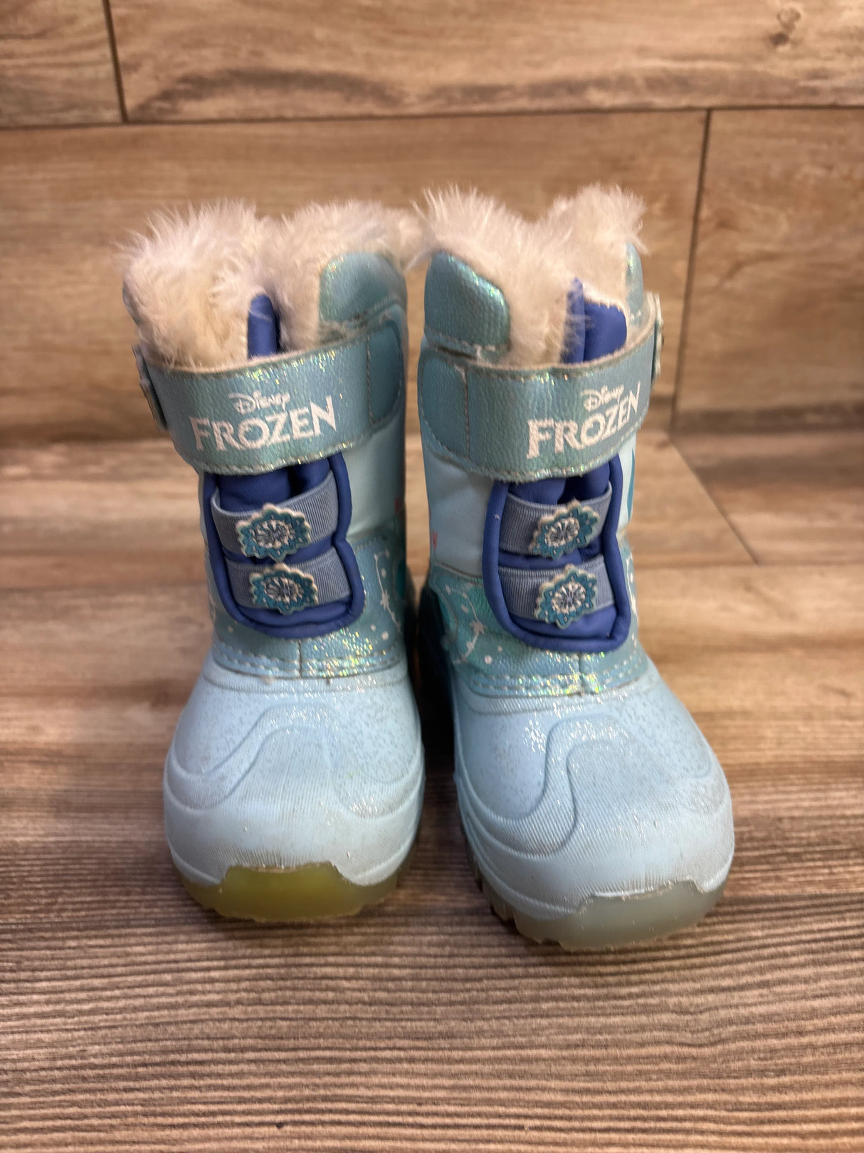 Front view of blue Disney Frozen light-up snow boots showing faux-fur cuff, snowflake fasteners and logo strap on wood floor