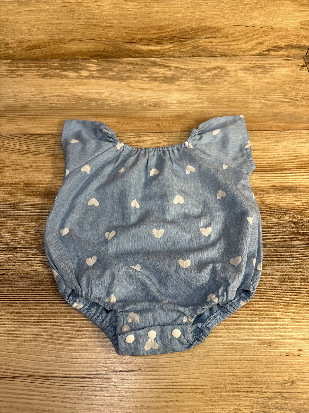 Top-down view of blue heart-print baby romper (size 3M) showing cap sleeves and snap crotch on wood background