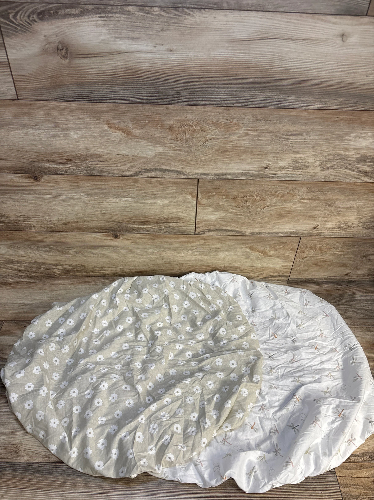 Two fitted crib sheets, one with white daisies on beige, one with dragonflies on white, on wood floor