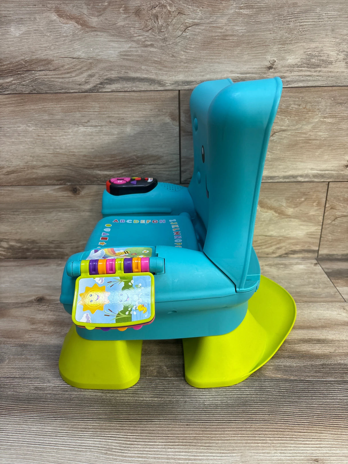 Side view of Fisher Price Laugh and Learn chair in teal showing yellow rocker base, flip toy and button panel on wood background