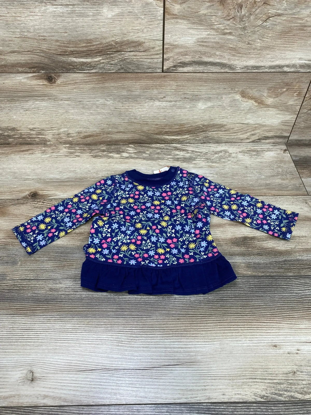 Flat lay of NEW Cat & Jack Floral Peplum Adaptive Shirt size 3-6m showcasing navy long-sleeve top with multicolor floral print and ruffle hem on wood plank floor