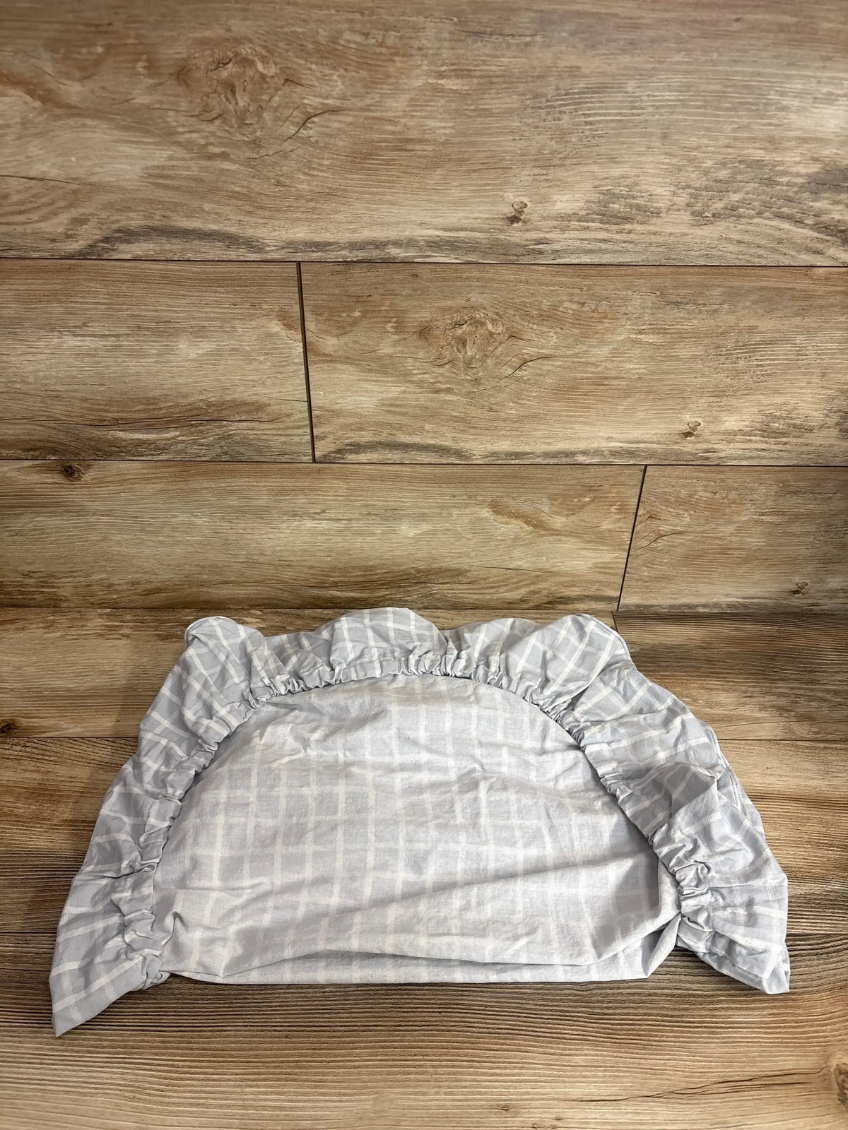 Top-down view of fitted bassinet sheet in light grey windowpane check showcasing gathered elastic edge with wood-plank background