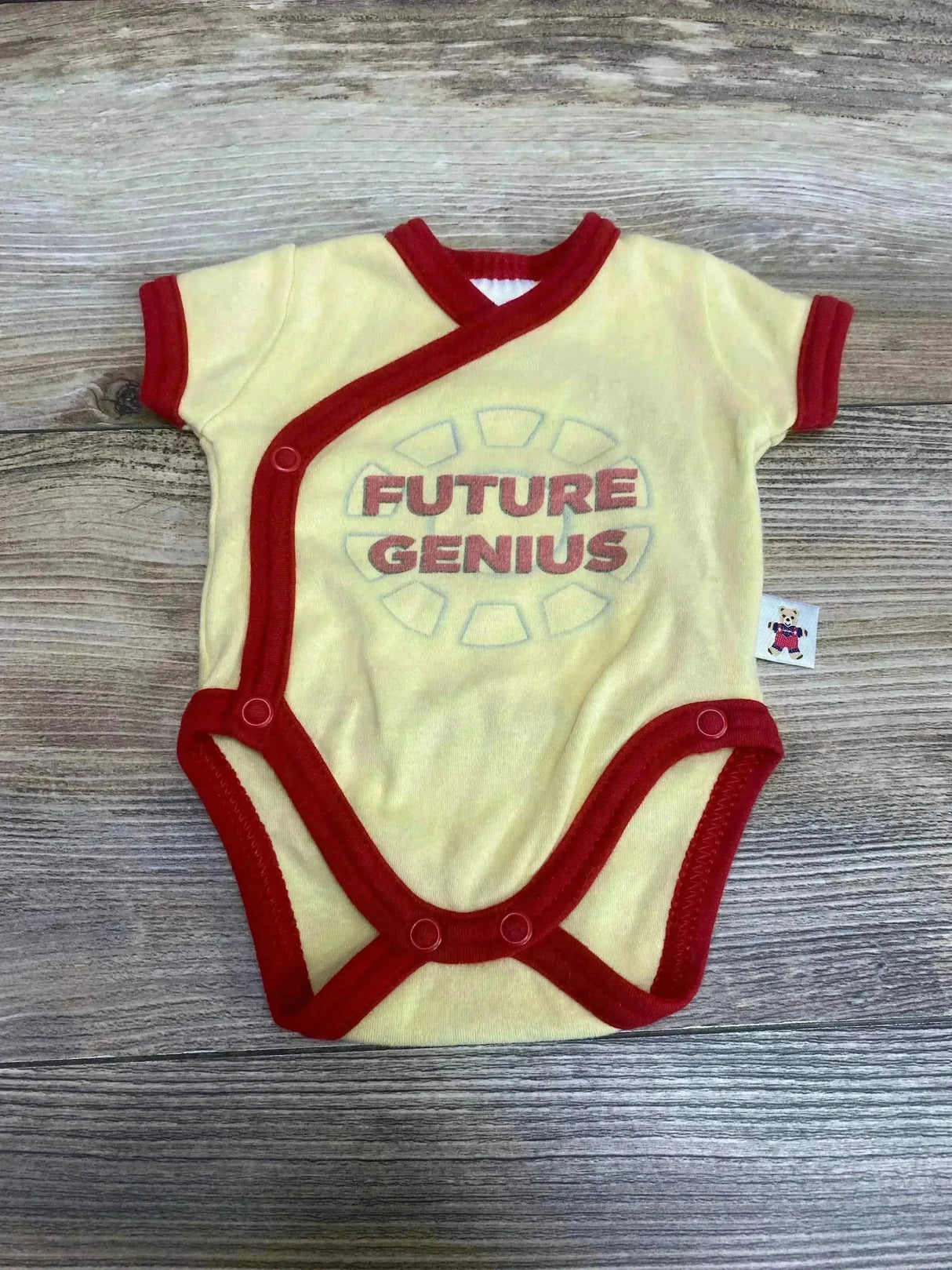 Flat-lay of preemie yellow kimono bodysuit with red trim showing FUTURE GENIUS text on wood floor background