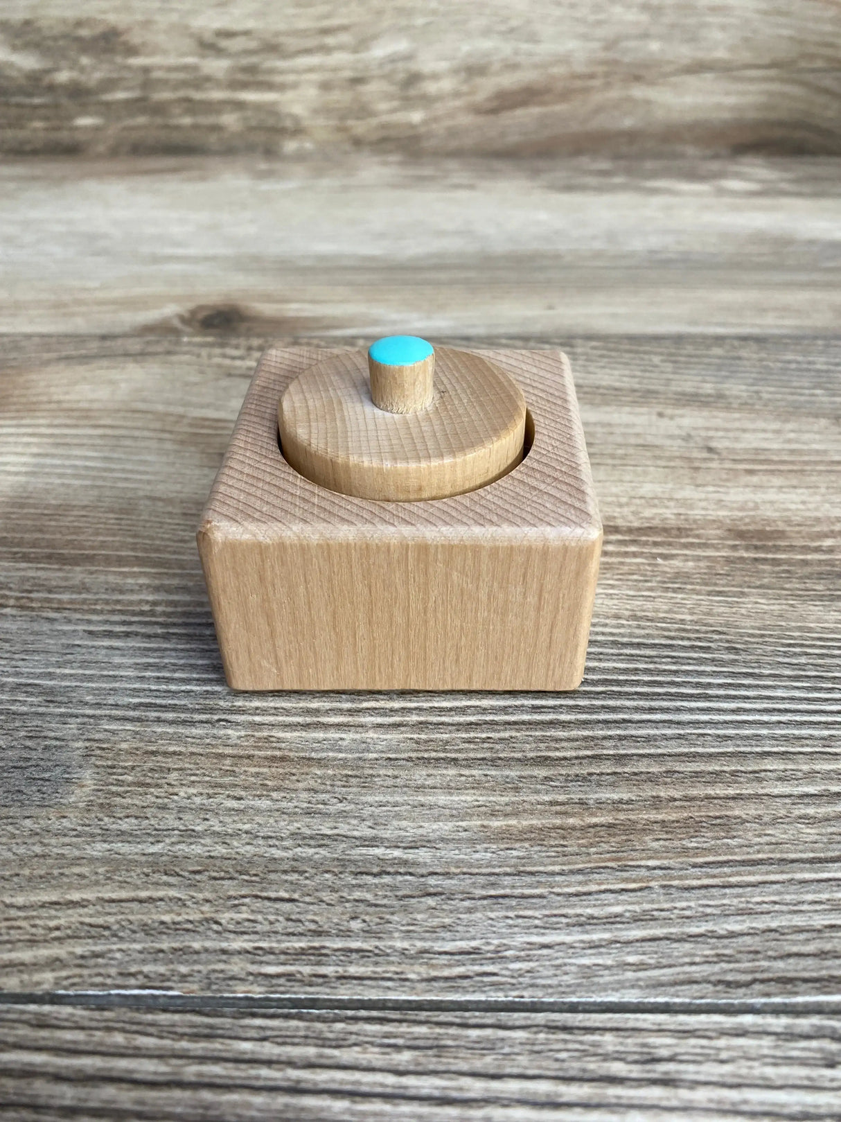Front view of Lovevery Pincer Puzzle showing natural wood block with circular lid and small turquoise knob on wood background