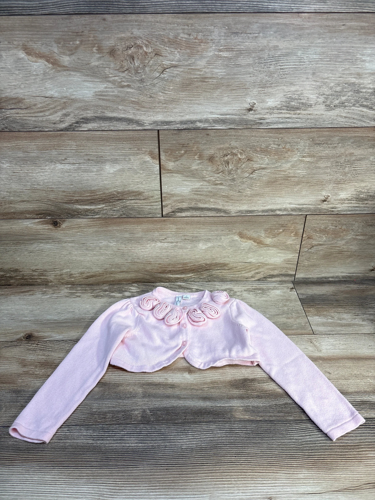 Light pink long sleeve bolero for girls with rosette trim, laid flat on wood background
