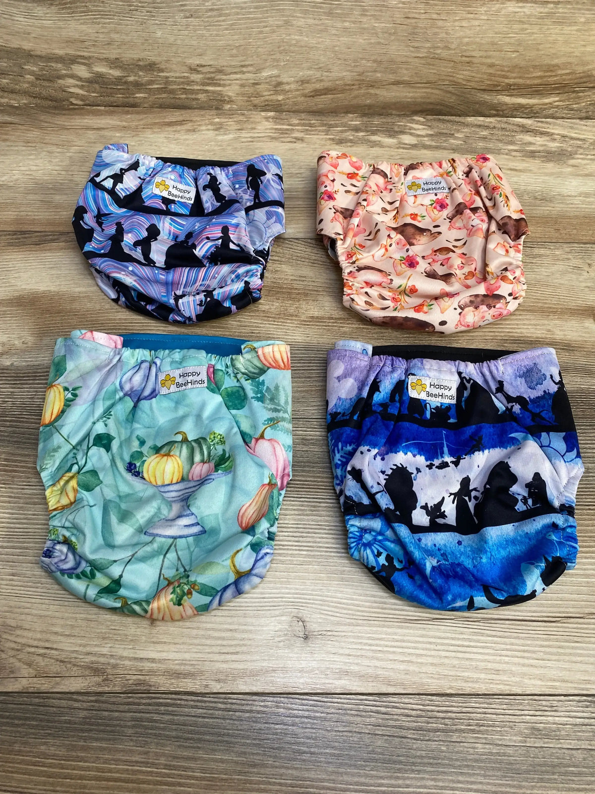 Flat lay of Happy BeeHinds 4Pk Pocket Diapers in four assorted prints purple silhouette pink floral teal pumpkin and blue fairy on wood plank floor