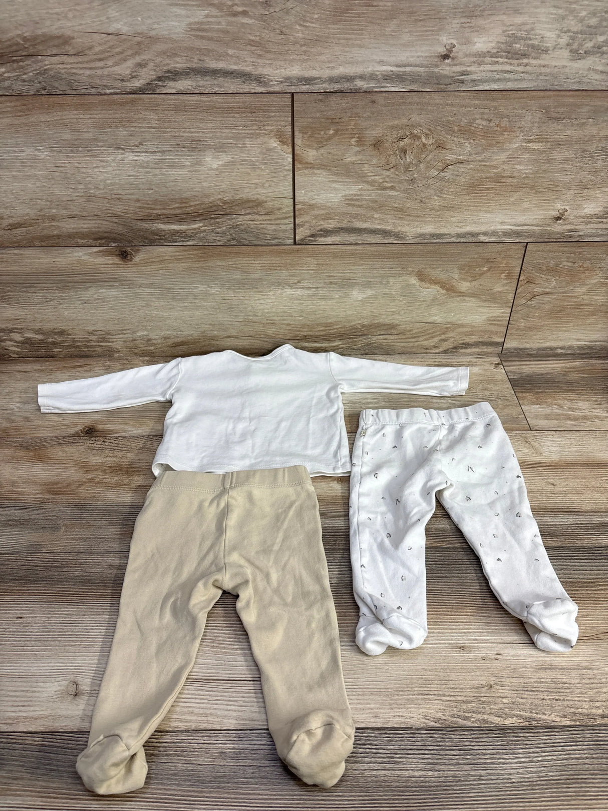 Baby clothing set with white long sleeve shirt, beige footed pants, and white printed footed pants on wood background