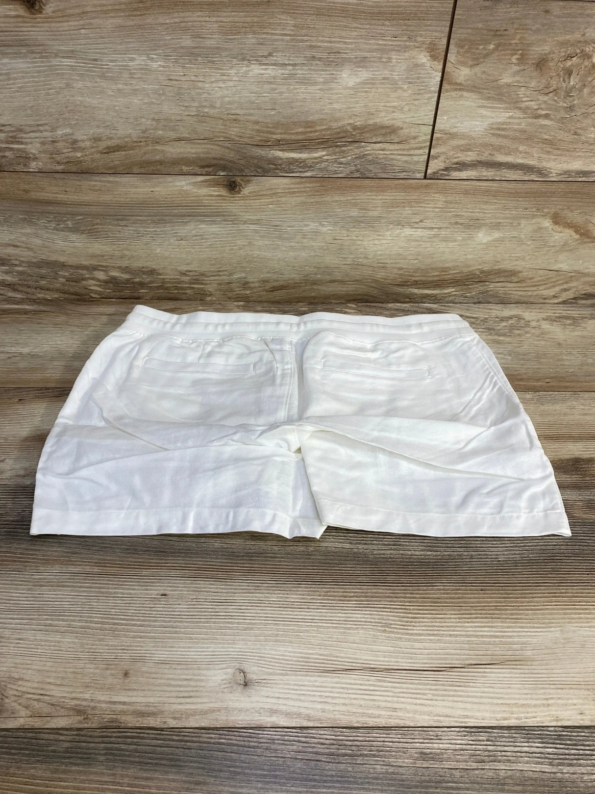 Flat lay of Gap maternity white linen-cotton shorts showing back pockets and elastic maternity waistband on wood plank background