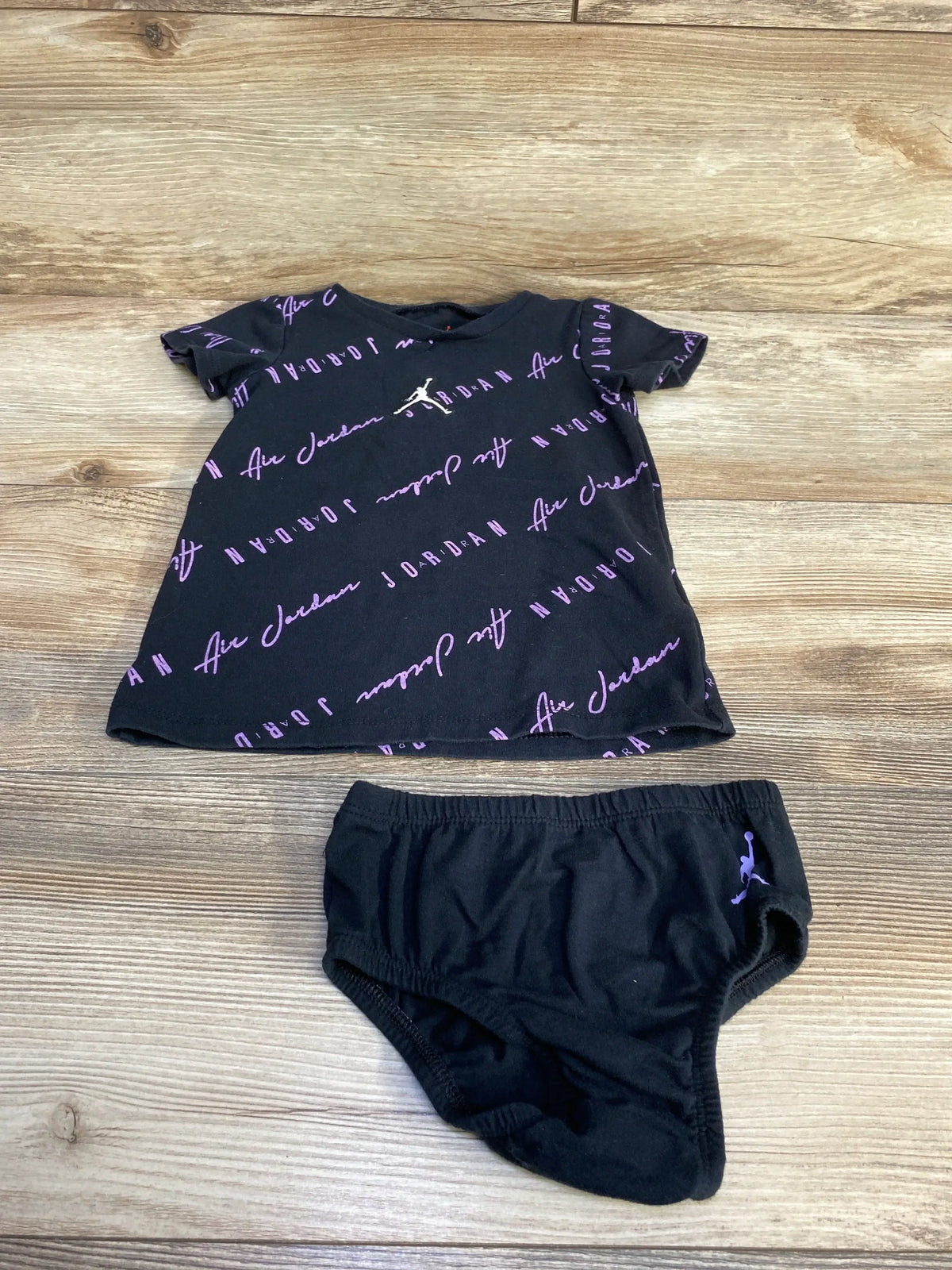 Flat lay of black baby dress and bloomers with purple diagonal Air Jordan script and Jumpman logos on wood background