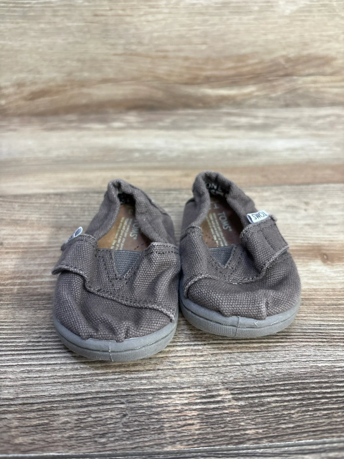 Front view of grey TOMS Tiny Alpargata toddler slip-ons showing elastic V-top, Velcro strap and worn rubber soles on wood surface