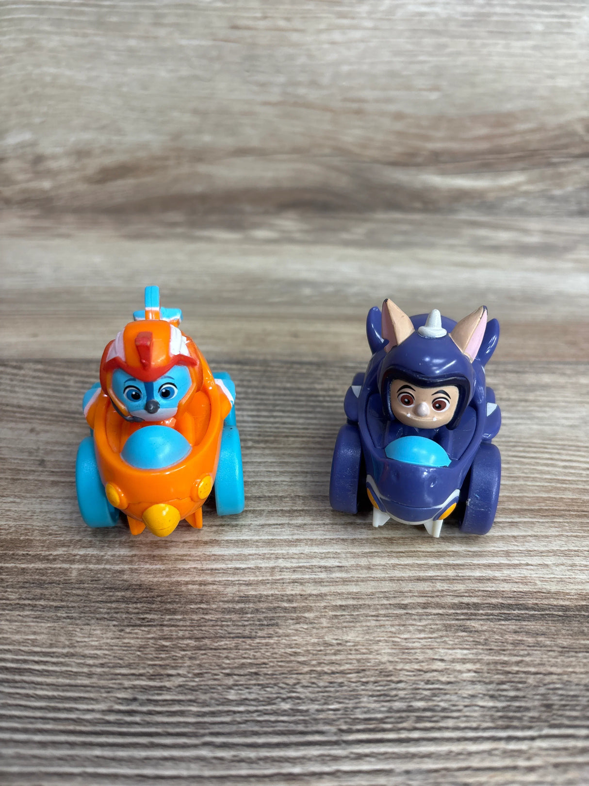 Two small children's toy cars on wooden surface, one orange and blue with a character, one purple with ears and face detail