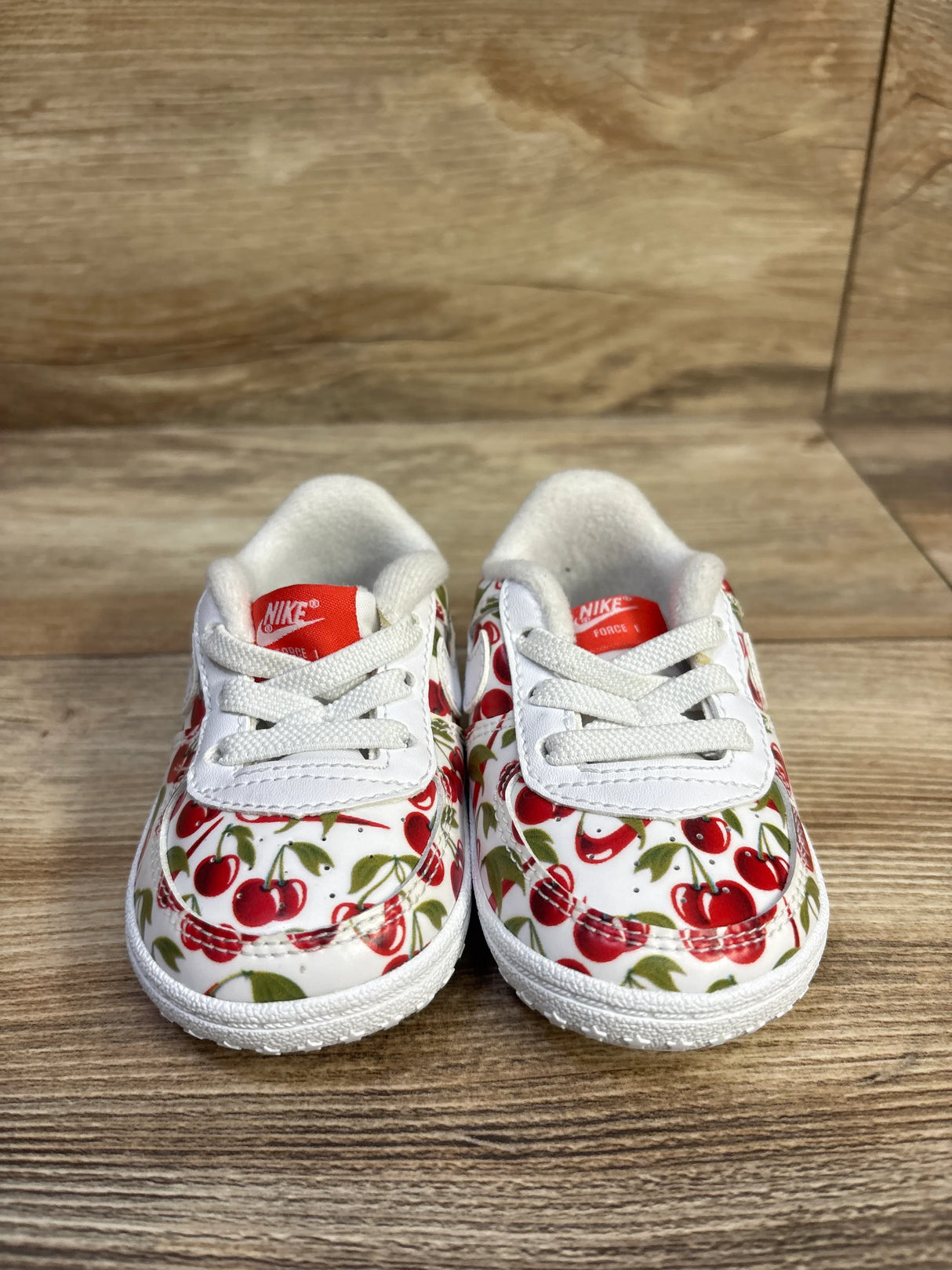 Front view of Nike Air Force crib shoes showcasing red cherry print and red Nike tongue label on a wood surface