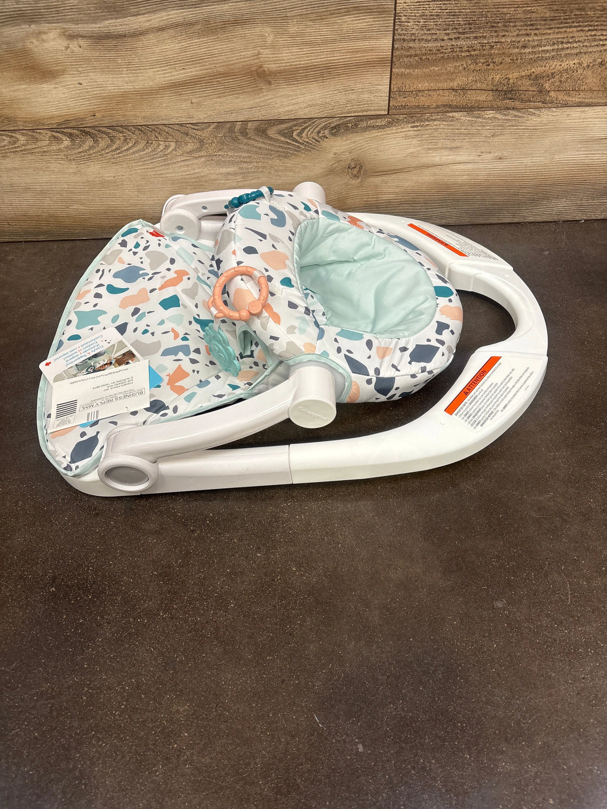 Folded white baby activity seat with blue and peach patterned fabric and attached toys on brown floor